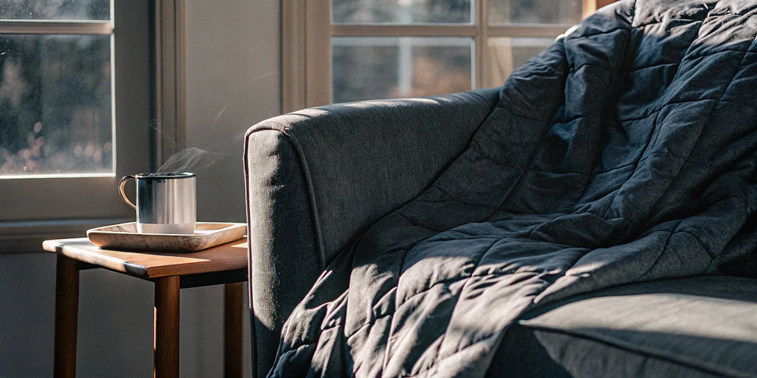 A gray deep pressure therapy blanket draped on a sofa next to a warm mug.