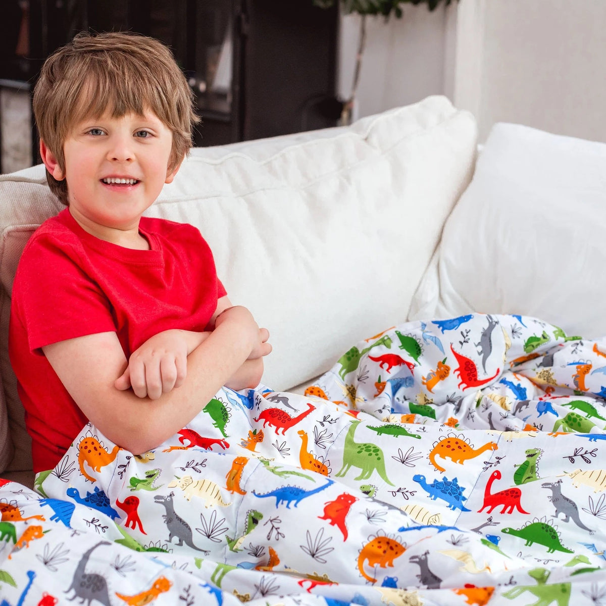 Little boy snuggled under his Mosaic Weighted Blankets Dinosaur Weighted Blanket in bed. 