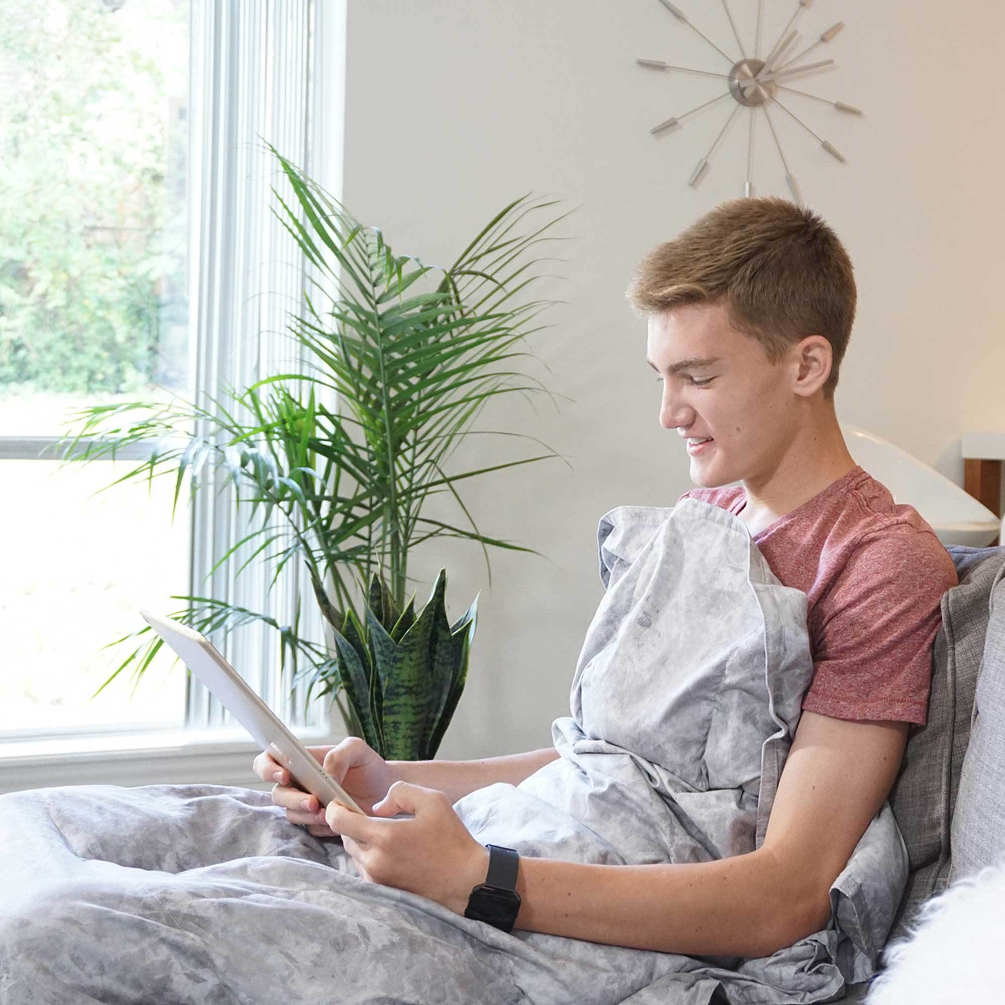 Man reads from his iPad snuggled under her Gray Cotton Weighted Blanket in bed