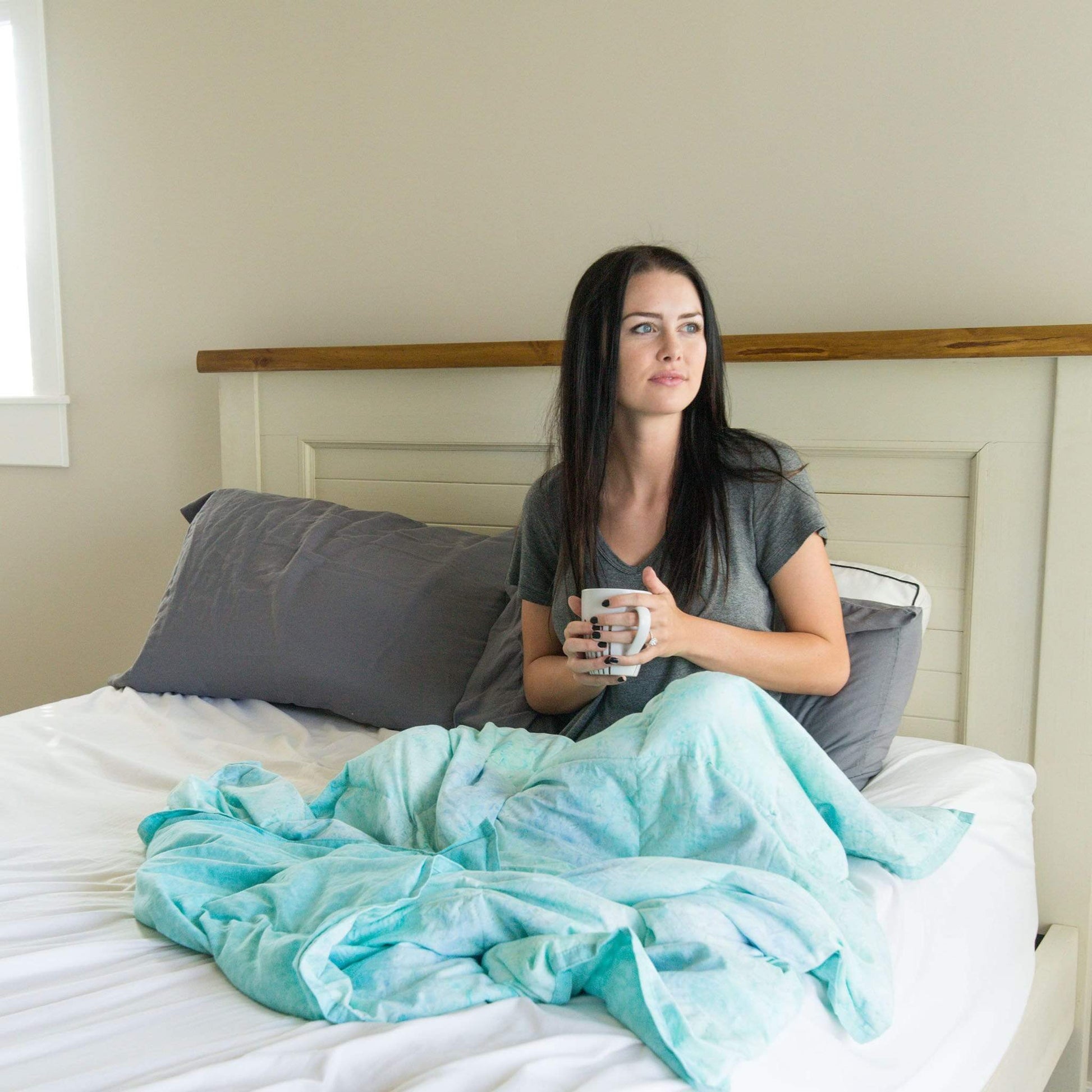 Woman snuggles and drinks from a mug under her Light Aqua Weighted Blanket on the couch in bed