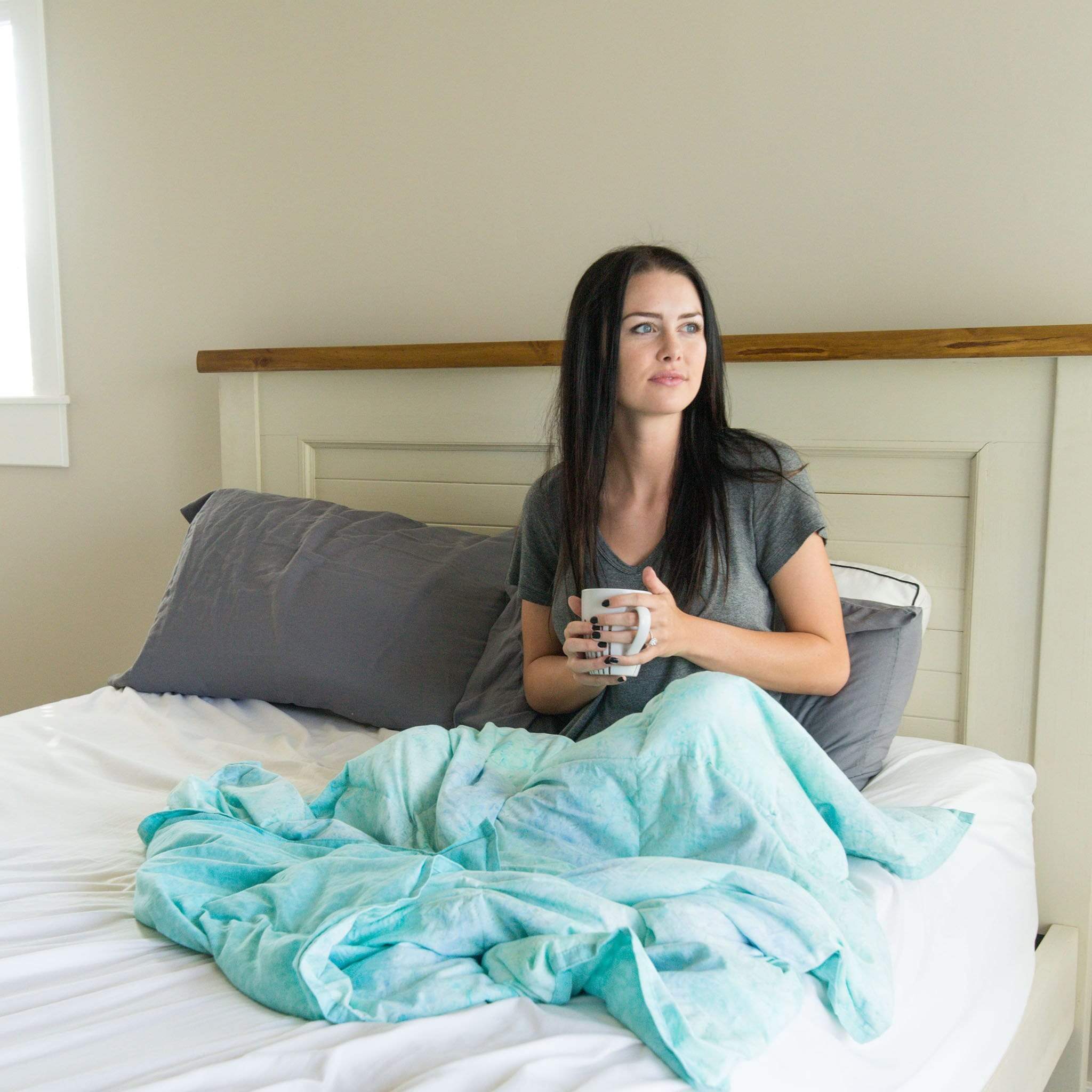 Woman snuggles and drinks from a mug under her Light Aqua Weighted Blanket on the couch in bed
